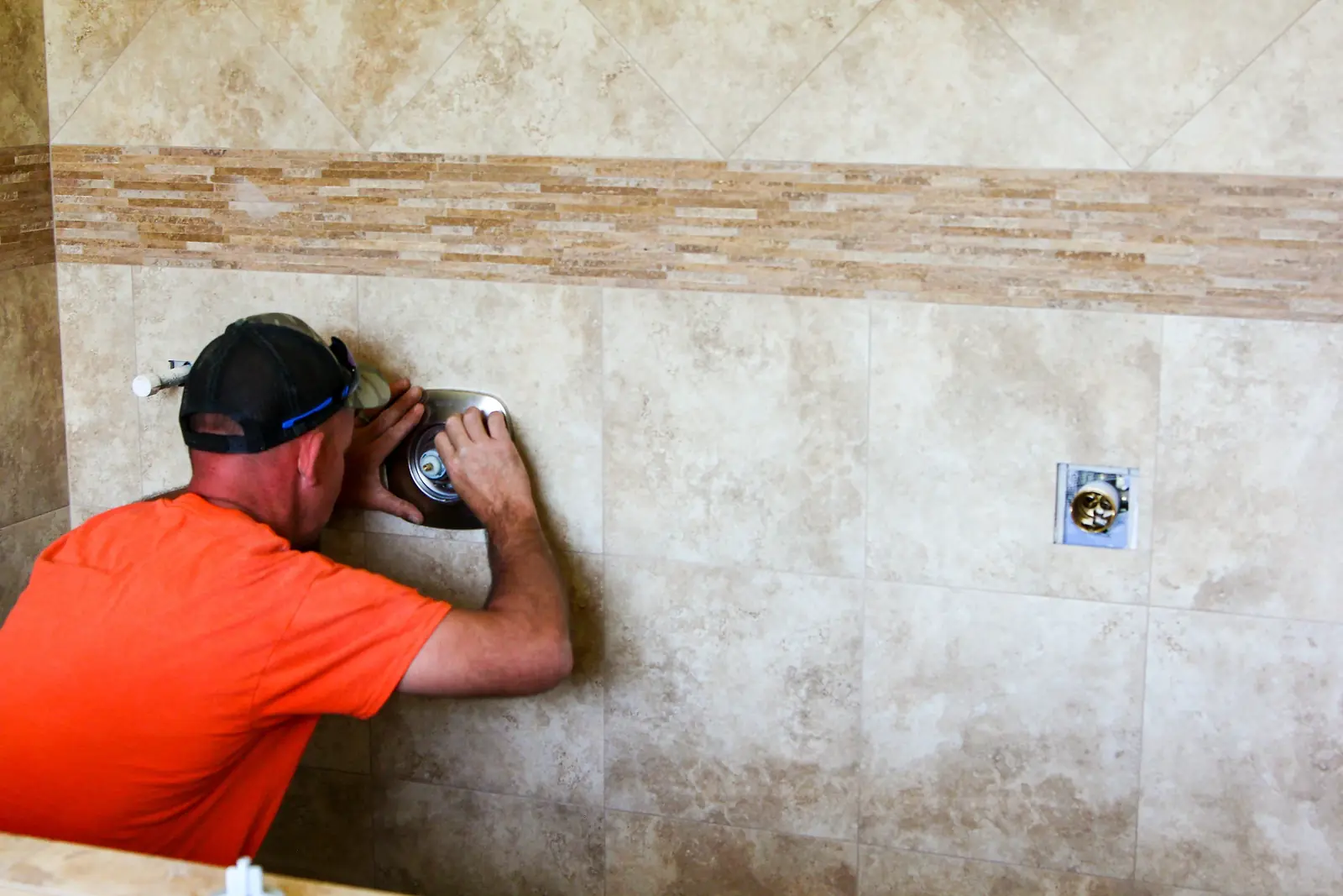 Plumber in an orange shirt fitting a shower valve trim plate to travertine tile