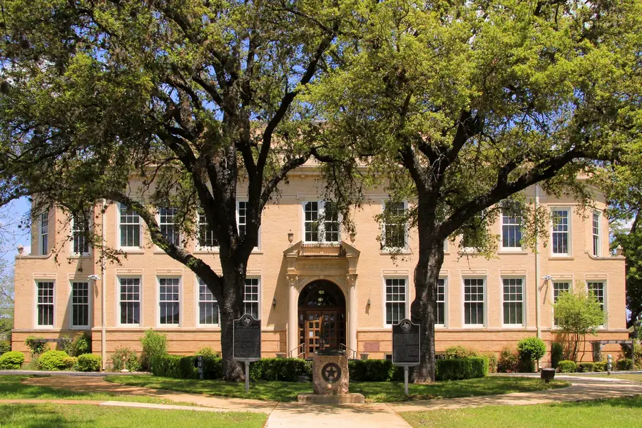 The Kerr County Courthouse in Kerrville, a tan brick building behind two big oak trees, with two state historical markers on the lawn