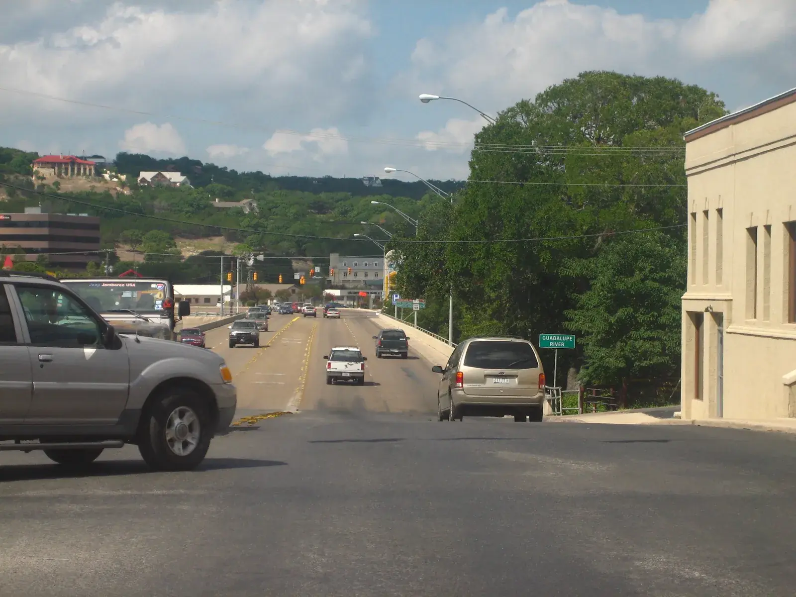 Cars on the bridge over the Guadalupe River into downtown Kerrville, a Guadalupe River sign at the rail and homes on the green ridge behind