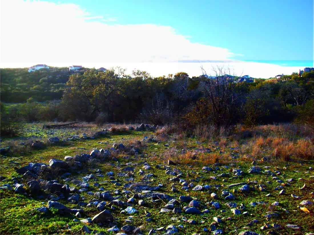 A field of loose limestone rock at Stone Oak Park in North San Antonio, with the rooflines of hillside homes on the far tree line